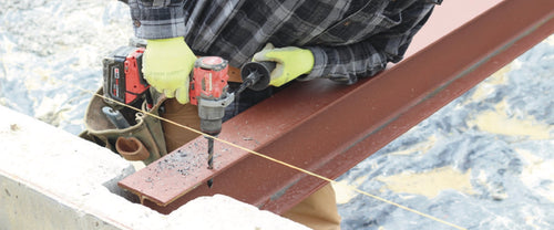 Worker drilling a steel I-beam flange with a cordless drill, metal chips on the beam