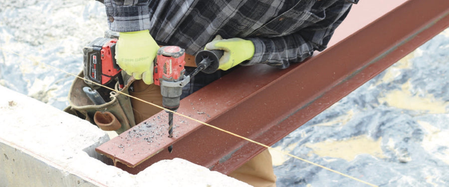 Worker drilling a steel I-beam flange with a cordless drill, metal chips on the beam