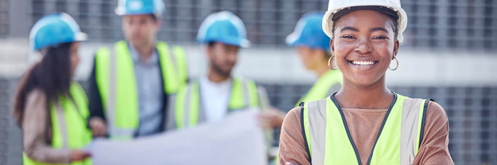 Construction manager in a hard hat and safety vest smiling, crew reviewing drawings behind her