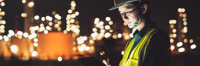 Civil engineer in a hard hat and safety vest reviewing plans on a tablet at night, plant lights behind