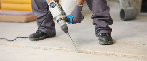 A construction worker in grey work pants and gloves bends over a concrete floor, chiseling a channel with a blue-and-white rotary hammer.